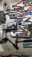 Photo showing assorted hand tools including hammers, pliers, screwdrivers, files, a staple gun, and clamps laid out on a workbench.