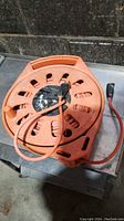 Photo of the orange extension cord wound on an orange plastic reel with a handle, showing plug and socket ends.