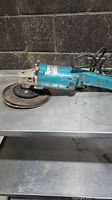Side view of blue Makita disc grinder with abrasive disc mounted showing overall wear and dirt.