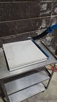 Manual paper slicer on metal cart against a wall, showing 16x16 inch grid cutting board and attached handle.