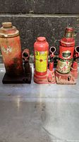 Photo shows three used hydraulic bottle jacks placed on a metal surface against a concrete wall. Left jack is large and rusty with missing label, middle jack is red with yellow 3-ton label, right jack is red with a 4-ton label and some wear.