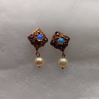 Close-up of the front of the earrings showing the square ornate gold setting with blue iridescent opal and dangling pearl on each earring.
