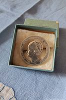 Top view of the sterling silver coin brooch inside a greenish-blue jewelry box with beige velvet lining.