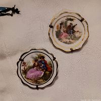 Two circular hand-painted Limoges porcelain brooches with romantic scenes and gold trim on a white fabric background.