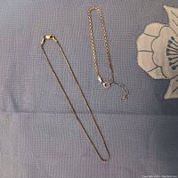 Two braided bracelets lying flat on a gray fabric surface, one gold and one silver in color, showing full length and clasp.