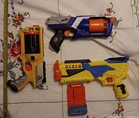 Three NERF toy guns laid out on a white tablecloth with a measuring tape beside the yellow and gray revolver-style blaster showing about 12-inch length.
