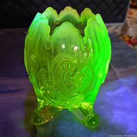 Front and slight side view of the green Vaseline glass vase showing its scalloped rim, embossed decorative floral/paisley pattern, and three-footed base. The photo shows the vase glowing under UV light.