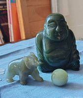 Close-up of dark green jade Buddha, light green jade elephant, and jade sphere on blue fabric.