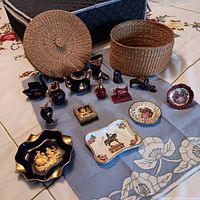 Woven basket with lid beside porcelain miniatures and ashtrays arranged on floral cloth