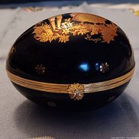 Closed egg-shaped Limoges box showing gold flower clasp and floral designs on a black background.