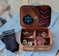 Open cariboo hide Kenwick jewelry box showing interior compartments filled with vintage cufflinks, pins, cloth patches, and badges including an RN badge and red chevrons.