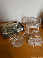 Wide view of candlesticks, votive candleholders in box, star-shaped dishes, ribbed bowl and block textured crystal piece on wooden surface against wall.
