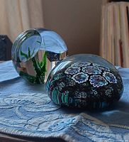 Photo of two glass paperweights displayed on blue embroidered fabric. One is clear with green plant element, the other is dark with floral millefiori pattern.
