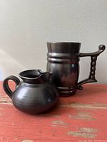 Jug and mug/stein displayed side by side on a red surface showing their shapes and gunmetal glaze