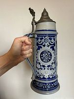 Stein held by the handle showing front floral patterns and pewter lid.