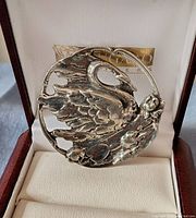 Close-up photo of silver-toned swan brooch in jewelry box showing detailed wing and floral design, tarnished patina.