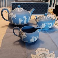 Photo of the full tea set showing the teapot, covered sugar dish and creamer on a table with detailed white relief against blue jasperware background.