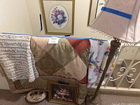 Photo showing standing lamp with pinkish decorative lampshade, quilted blanket with patchwork design, fleece blanket with colorful floral print, ceramic bed warmer pot, and framed floral wall art in background.