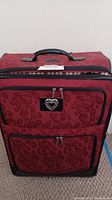 Front view of the burgundy Brighton luggage showing floral print, front compartments, and decorative heart-shaped zipper pull.