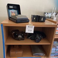 Overview of lot on wooden shelving: top shelf holds Polaroid Spectra System camera in case and Nikon flash, middle shelf holds Zeiss Ikon folding camera in case and Nikon film SLR, bottom shelf holds wooden shoe shine box and metal flask.