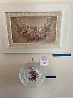 Floral painting with ornate white frame mounted above a floral porcelain hanging plate with Maxsold lot tag.