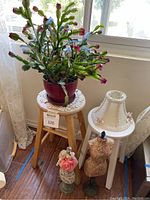 Full view showing live Christmas cactus plant on light wood stool with white wooden stool next to it holding cream-colored lamp shade and two miniature dress forms on the floor.