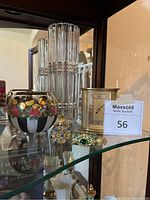 Photo showing tall clear cut glass vase, round hand-painted vase, gold-colored clock, and two small bejeweled perfume bottles on glass shelf.