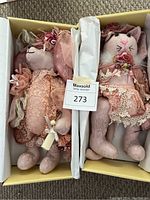 Two plush animals side by side in a box, one a teddy bear and the other a cat, both wearing pink lace outfits and floral hats.