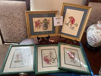Five framed frog needlepoint artworks on a table, two with gold frames standing on stands and three with green frames laid down