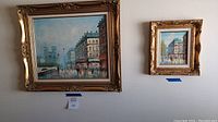 Front view of two framed city walk paintings on a wall, large and small, with blue tape markers below.