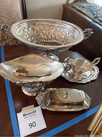 Photo showing all four silver-like serving pieces on wooden surface: large ornate tureen, pedestal bowl, gravy boat with tray, butter dish with lid.