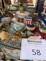 Overview of assorted porcelain and enamel trinket boxes arranged on glass surface showing various shapes and floral designs.