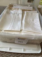 Clear plastic bin containing folded white linens with delicate embroidery and lace along edges.