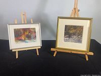 Two framed watercolour paintings on small easels. Left painting shows colorful trees and water, right painting shows a rocky river scene.