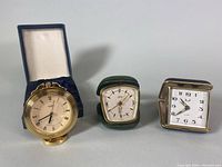 Front view of three clocks: brass Henry Birks clock in original box, green leatherette clock, and gold-tone folding clock.
