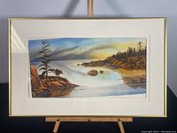 Framed watercolor painting showing a coastal landscape with trees, rocks, water, and sky, with the artwork slipped inside the frame under glass.