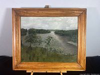 Framed landscape oil painting showing a river or beach scene with trees and cloudy sky, medium brown wooden frame.