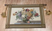Full view of the large vintage handmade floral tapestry laid flat on floor, showing a bouquet of flowers and ornate border with rod and tassels at the edges.