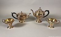 Full view of all four pieces: two teapots, creamer, and sugar bowl showing silver plated embossed decoration and black handles.