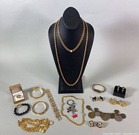 Full lot of gold-tone vintage costume jewelry including rings, bracelets, necklaces, earrings, and keychain, arranged on gray surface with black necklace display bust