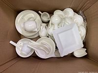 Top view of assorted white bone china dishes in a cardboard box showing small plates, bowls, pitchers, and ladles.