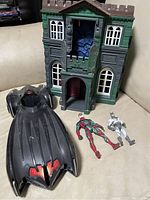 Photo shows a Batman themed playset building facade, a black Batmobile with red interior, and two action figures lying on the surface.