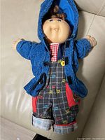 Cabbage Patch Kid doll wearing a blue knit hoodie, red checkered shirt, and checkered overalls with colorful CPK logos, laying on a beige surface