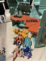 Castle Grayskull playset with orange platforms and green tower, four action figures in front, several yellow accessories on couch