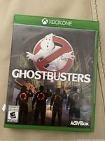 Xbox One game case for Ghostbusters laid on a beige surface. The cover shows four Ghostbusters characters and the Ghostbusters logo.