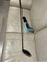 Full view of golf driver club with its head cover placed on a beige leather couch, showing the club's shaft and grip.