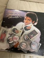 Photo of Michael Jackson Thriller vinyl record with visible cover damage and multiple sealed collectible pins and keychains featuring Michael Jackson images.