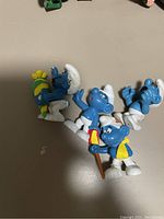 Four blue Smurf figurines with white pants and hats on a table, one holding a walking cane