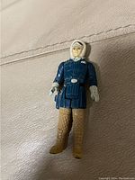 Photo shows vintage Han Solo Star Wars figure wearing a dark blue jacket, white gloves, tan quilted pants, and brown boots with a white hood on the head.