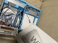 Photo showing Nintendo Wii console, white Wii Remote controller, sensor bar, power and AV cables, and a stack of seven Wii games including Just Dance 2014 and Sonic Racing.